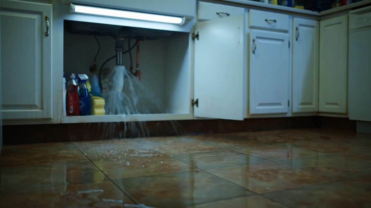 Pipe under a kitchen sink spraying water across a tile floor