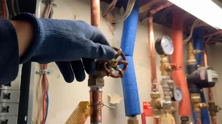 Gloved hand closing a red main water shut-off valve on copper plumbing in Anchorage, AK